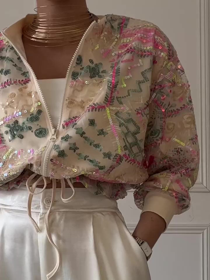 Woman wearing an apricot cropped bomber jacket with multicolored sequin embroidery over white trousers.